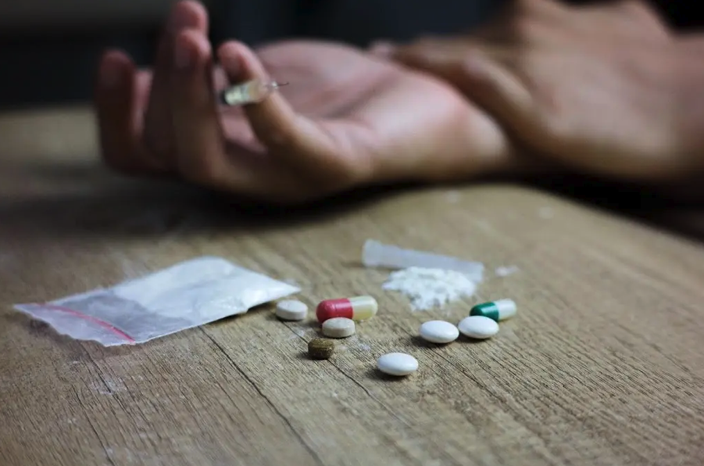Various pills, a small plastic bag with white powder, a pile of white powder, and a syringe in the background held by a hand